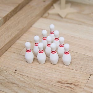 Könnte beinhalten: Ein Miniatur-Bowling-Set mit weißen Kegeln mit roten Streifen und einem kleinen Ball mit bunten Tupfen. Das Set ist auf einer hellen Holzoberfläche angeordnet, was auf ein Spiel oder einen Dekorationsartikel hindeutet.