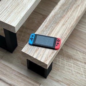 Könnte beinhalten: Eine tragbare Miniatur-Spielkonsole mit schwarzem Bildschirm, blauem linken Controller und rotem rechten Controller, die auf einer kleinen Holzbank mit schwarzen Beinen liegt. Die Bank ist Teil eines Miniatur-Picknicktisch-Sets aus hellem Holz.