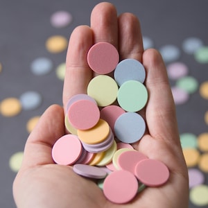 Könnte beinhalten: Eine Hand hält einen Haufen runder Konfetti in Pastellfarben. Das Konfetti ist in den Farben Rosa, Gelb, Blau und Grün.