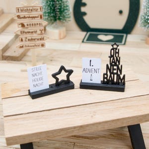 Könnte beinhalten: Zwei schwarze rechteckige Ständer mit weißen Karten und schwarzen Dekoelementen. Eine Karte zeigt STILLE NACHT. HEILIGE NACHT mit einem Stern, die andere 1. ADVENT mit Kerzensymbol und einem schwarzen OH TANNENBAUM-Schriftzug mit Stern.
