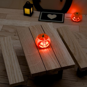Könnte beinhalten: Ein Miniatur-Picknicktisch aus Holz mit befestigten Bänken, auf dem ein kleiner, leuchtend orangefarbener Kürbis mit geschnitztem Gesicht steht. Die Szene enthält eine winzige schwarze Laterne und einen weiteren kleinen leuchtenden Kürbis.