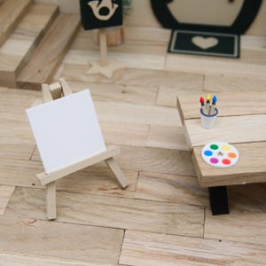 Könnte beinhalten: Miniatur-Kunstbedarf-Set, einschließlich einer kleinen Holzstaffelei mit einer leeren weißen Leinwand, einem Holztisch mit einer Farbpalette und einem kleinen Becher mit bunten Pinseln. Die Szene spielt auf einem Holzboden.