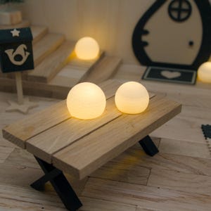 Könnte beinhalten: Zwei kleine, kugelförmige, leuchtend weiße LED-Lichter mit strukturierter Oberfläche stehen auf einem Miniatur-Picknicktisch aus Holz. Der Tisch besteht aus hellen Holzplanken und schwarzen, kreuzförmigen Beinen und sorgt für warmes Licht.