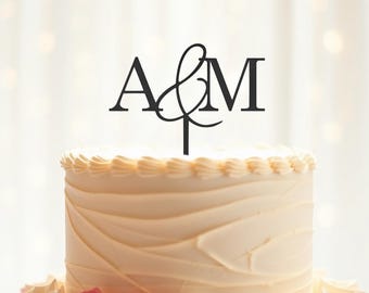 以下が含まれることがあります： 白いウェディングケーキに、黒いケーキトッパーが乗っています。ケーキトッパーには、筆記体で「A&M」と書いてあります。