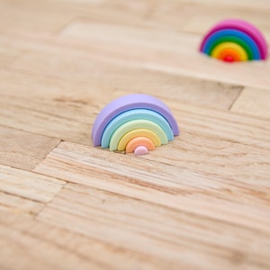 Op de afbeelding: Twee regenboogvormige objecten bestaande uit gestapelde halfronde bogen. Eén object heeft pasteltinten van paars, blauw, groen, geel en perzik. Het tweede object op de achtergrond toont levendige kleuren van roze, blauw, groen en rood.