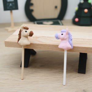 Könnte beinhalten: Zwei Miniatur-Pferdekopf-Figuren auf Stäbchen. Ein Pferd ist beige mit brauner Mähne und Schnauze, das andere ist hellrosa mit lavendelfarbener Mähne und Schnauze. Beide haben schwarze Augen und sind auf dünnen, geraden, vertikalen Stäben befestigt.