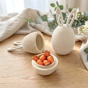 Könnte beinhalten: Behälter in Form eines Osterhasen aus cremefarbener Keramik mit vertikalem Rippendesign. Der Behälter hat einen Deckel und eine kleine Schale, die mit orangefarbenen, folienverpackten Süßigkeiten gefüllt ist. Ein Behälter ist geöffnet, ein anderer liegt auf der Seite.