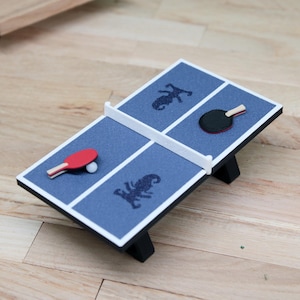 Könnte beinhalten: Ein blauer Miniatur-Tischtennistisch mit weißen Begrenzungslinien und einem weißen Netz in der Mitte. Die Oberfläche zeigt zwei dunkelblaue Seepferdchen-Silhouetten. Eine Seite hat einen roten Schläger und einen weißen Ball, die andere einen schwarzen Schläger.