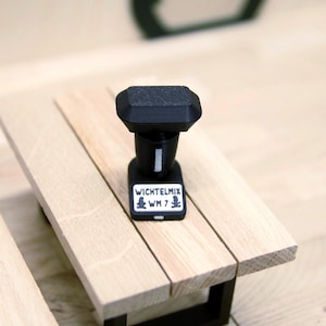 Könnte beinhalten: Ein schwarzes Miniatur-Objekt aus Kunststoff mit rechteckiger Oberseite und einem Sockel, der ein weißes Etikett mit dem Text WICHTELMIX WM 7 sowie zwei kleine schwarze Silhouetten-Figuren auf beiden Seiten trägt, platziert auf einer hellen Holzfläche.