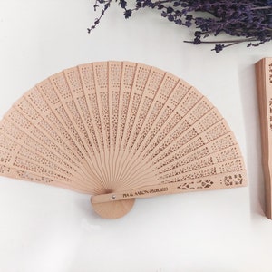 May include: Two wooden hand fans with intricate cutouts. One fan is closed and the other is open. The closed fan has the text "VICTORIA & MARK" engraved on it. The open fan has the text "TIA & AARON 05.08.2023" engraved on it.