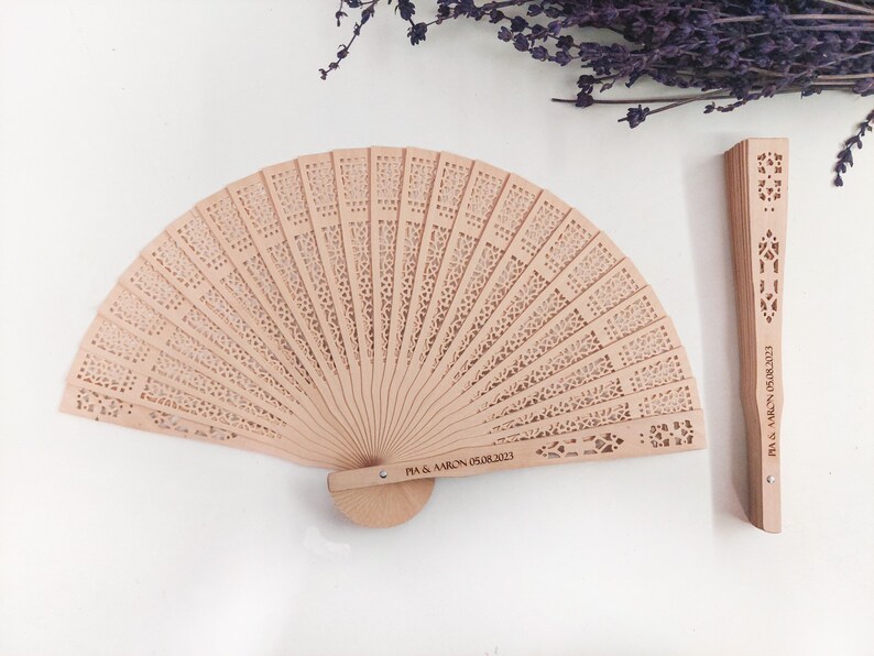 May include: Two wooden hand fans with intricate cutouts. One fan is closed and the other is open. The closed fan has the text "VICTORIA & MARK" engraved on it. The open fan has the text "TIA & AARON 05.08.2023" engraved on it.