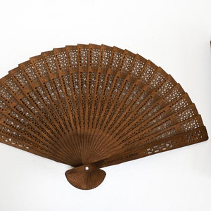 May include: A brown wooden folding fan with intricate cutouts and the text "A Moment Of 12.2023". The fan is closed and lying on a white surface. Next to it is a matching wooden fan stick.
