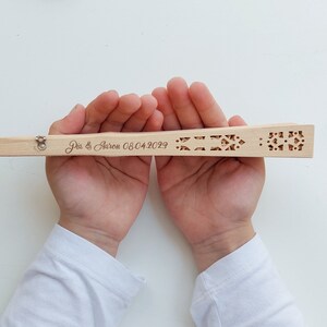 May include: A wooden hand fan with a decorative cutout design. The fan is personalised with the text "Pia & Aaron 08.04.2023".