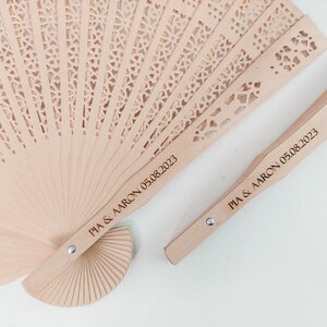 May include: Two wooden hand fans with a delicate, intricate design. The fans are personalised with the text "PIA & AARON 05.08.2023".