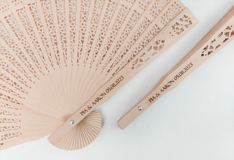 May include: Two wooden hand fans with a delicate, intricate design. The fans are personalised with the text "PIA & AARON 05.08.2023".