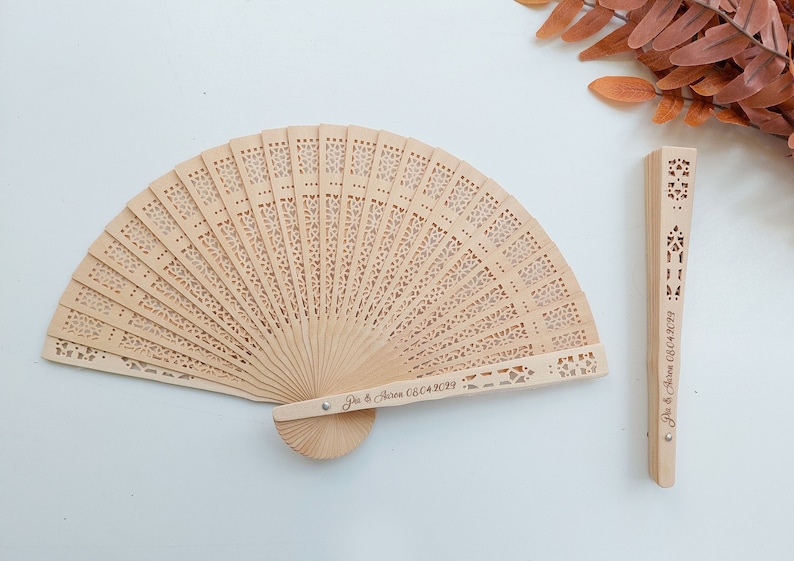 May include: A wooden hand fan with a delicate, intricate design. The fan is closed and has a personalised inscription that reads "Joe & Aaron 08.04.2023".