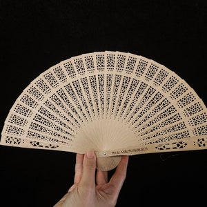 May include: A light brown wooden hand fan with a delicate, intricate design. The fan is closed and has a personalised inscription that reads "PIA & AARON 05.08.2023".