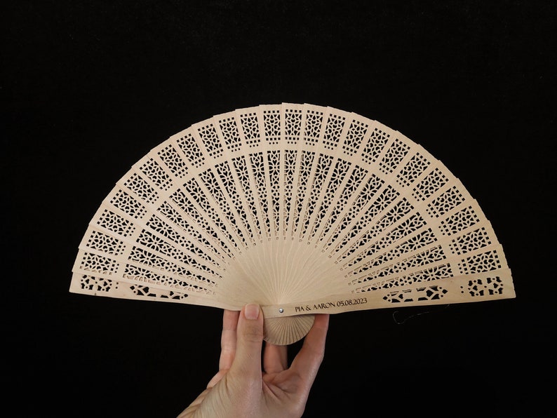 May include: A light brown wooden hand fan with a delicate, intricate design. The fan is closed and has a personalised inscription that reads "PIA & AARON 05.08.2023".