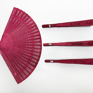 Puede incluir: Tres abanicos plegables de madera roja con intrincados diseños tallados. Los abanicos están cerrados y dispuestos horizontalmente sobre una superficie blanca.