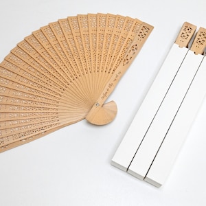 以下が含まれることがあります： 複雑な切り抜きと「Sofia & Lily 14.07.23」というパーソナライズされた刻印が施された、木製の折りたたみ式の扇子が3つセットになっています。扇子は白い段ボール箱に梱包されています。