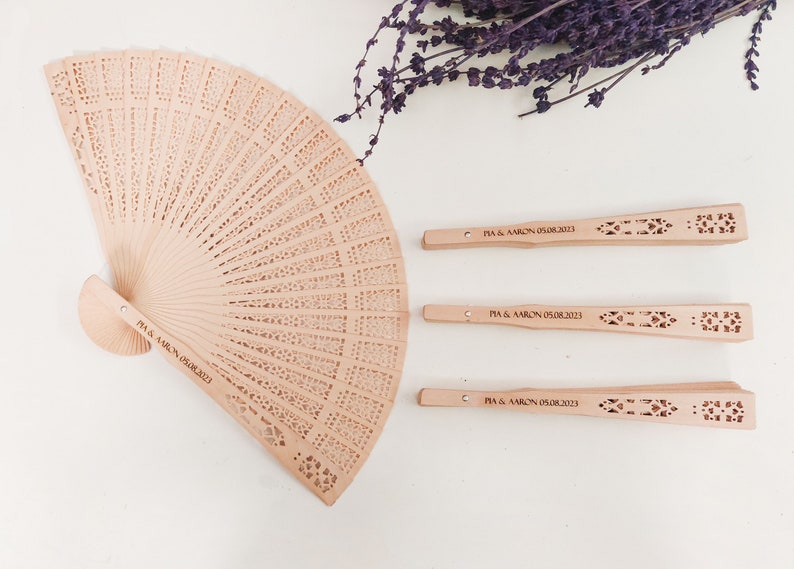 May include: Four wooden hand fans with a decorative cut-out design. The fans are personalised with the text "PIA & AARON 05.08.2013".