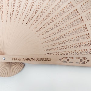 May include: A light brown wooden hand fan with a delicate, intricate cut-out design. The fan handle is engraved with the text "PIA & AARON 05.08.2023".