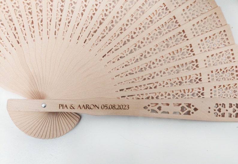 May include: A light brown wooden hand fan with a delicate, intricate cut-out design. The fan handle is engraved with the text "PIA & AARON 05.08.2023".
