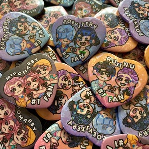 May include: A selection of heart-shaped fan art badges. Each badge displays vibrant cartoon illustrations of characters, accompanied by the text "#1 FAN" and names such as "ZOEYSTERY" and "MIROMABBY".