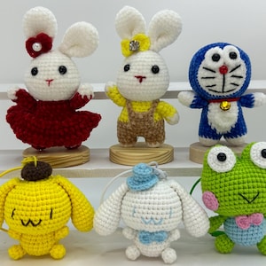 May include: A collection of six crocheted stuffed animals, including a yellow bunny with a brown overalls, a white bunny with a red dress, a blue cat with a bell, a green frog with a blue bow tie, a white dog with a blue bow tie, and a yellow dog with a brown hat.