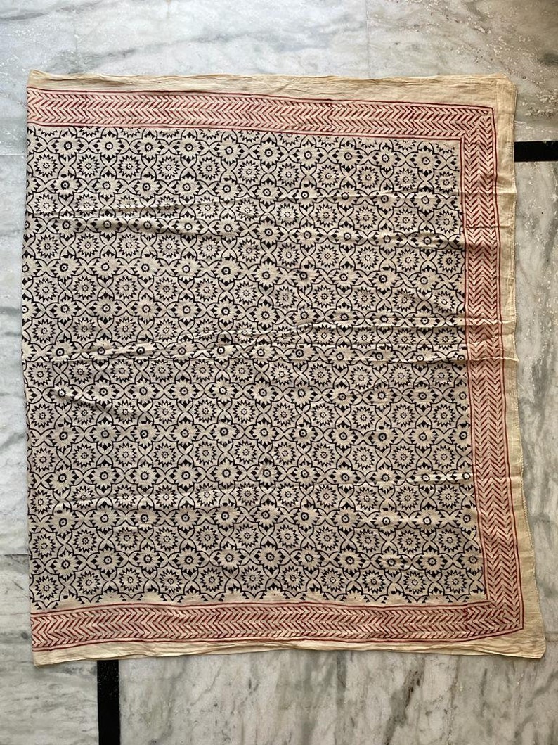 May include: A cream-colored cotton fabric with a black and white floral pattern. The fabric has a red border with a geometric design.