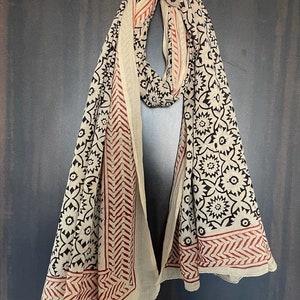 May include: A cream-colored cotton scarf with a black and red floral print. The scarf is folded and hanging on a hook.