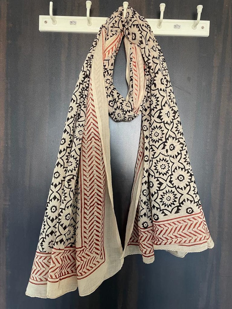 May include: A cream-colored cotton scarf with a black and red floral print. The scarf is folded and hanging on a hook.