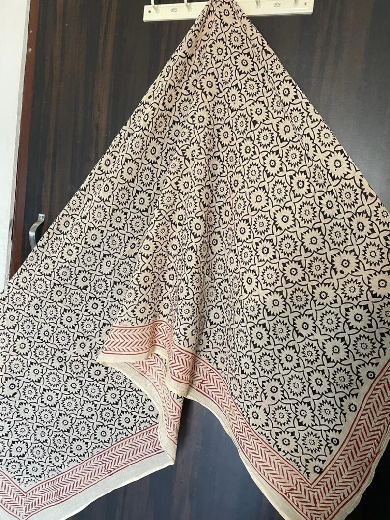 May include: A cream-colored cotton scarf with a black floral pattern and a red border. The scarf is folded and hanging on a hook.