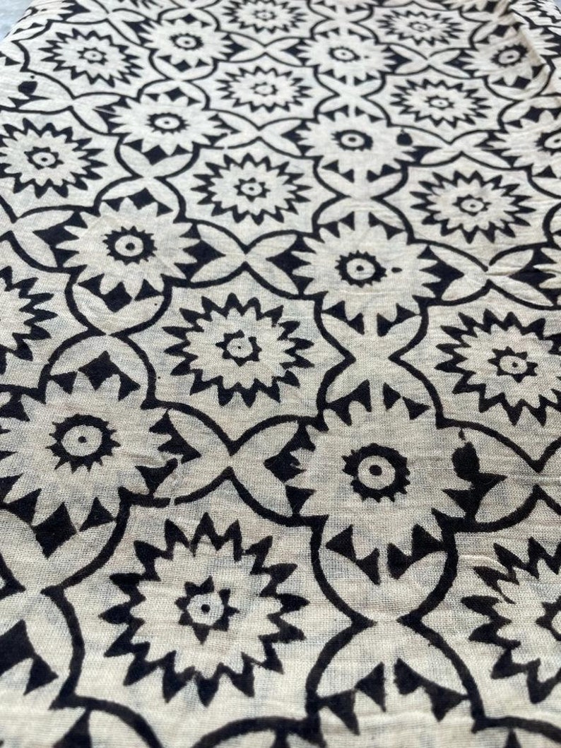 May include: Black and white fabric with a repeating geometric pattern of stylized flowers. The fabric has a slightly textured surface.