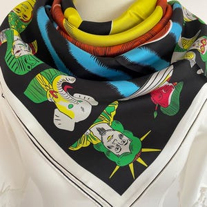 May include: A black silk scarf with a white border. The scarf features a colourful design of a woman's face, an elephant, and other figures. The scarf is folded and displayed on a white mannequin.