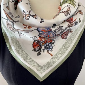 May include: A square, white silk scarf featuring a detailed butterfly and floral design. The scarf has a green border with a dotted pattern. The butterflies are in shades of orange, blue, and brown. The scarf is draped around a neck.