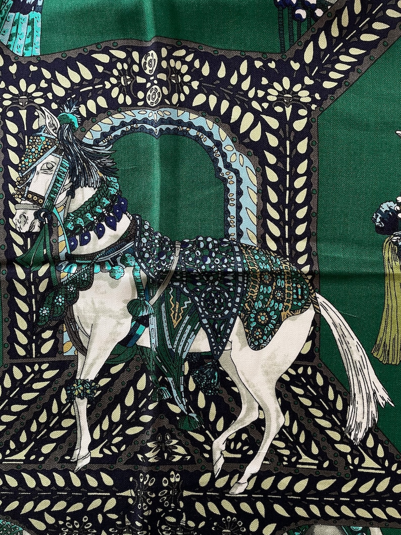 Peut inclure: Un foulard en soie vert avec une illustration d&eacute;taill&eacute;e d'un cheval blanc orn&eacute; d'une sellerie orn&eacute;e. Le cheval est plac&eacute; sur un fond d'arcades d&eacute;coratives et de feuillage dans des tons de vert, de bleu et de noir.