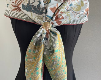May include: A white silk scarf with a colorful floral and animal print. The scarf is tied around the neck of a mannequin wearing a black dress.