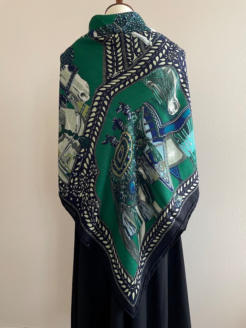 Peut inclure: Un foulard en soie vert avec un motif &eacute;quin. Le foulard pr&eacute;sente un fond vert fonc&eacute; avec des accents blancs, bleus et dor&eacute;s. La bordure est bleu marine fonc&eacute; avec un motif de feuilles blanches. Le foulard est drap&eacute; sur un mannequin noir.
