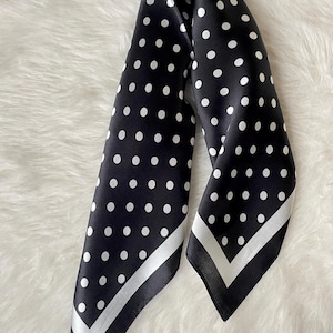 May include: A black and white polka dot scarf. The scarf is made of a silky material and features white polka dots on a black background. The edges are trimmed with a white and black border. The scarf is folded and draped.