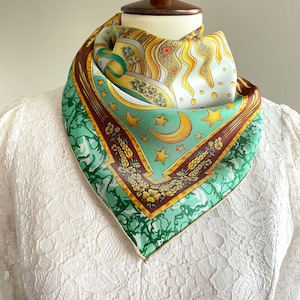 May include: A green, yellow, and brown silk scarf with a pattern of stars, a crescent moon, and a sun. The scarf is tied around a white lace mannequin.