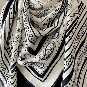 May include: A black and white patterned scarf. The scarf has a square shape and is decorated with paisley motifs, stripes, and other designs. The fabric is draped, highlighting the detailed pattern and texture of the material.