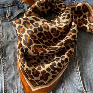 May include: A square scarf featuring a brown, black, and tan leopard print pattern. The scarf has a solid brown border with thin white and black stripes along the edges. The fabric has a smooth, satin-like finish and is folded into a triangle.