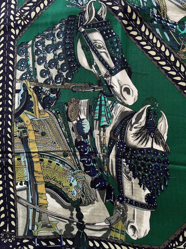 Peut inclure: Un carr&eacute; de soie avec un motif &eacute;questre d&eacute;taill&eacute;. L'&eacute;charpe pr&eacute;sente deux chevaux blancs avec des harnachements et des brides orn&eacute;s, sur un fond vert fonc&eacute;. Le motif comprend des &eacute;l&eacute;ments d&eacute;coratifs dans les tons de bleu, d'or et de vert.