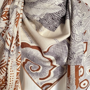 May include: A cream-colored scarf with intricate patterns in brown and gray. The scarf features detailed botanical and geometric designs, creating a visually rich textile. The scarf is draped, showcasing its flowing fabric and design.