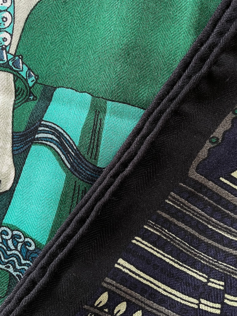Peut inclure: Gros plan sur un foulard &agrave; motifs avec une bordure bleu marine fonc&eacute;. Le foulard pr&eacute;sente un motif avec des nuances de vert, de sarcelle et de bleu marine. Le tissu a un aspect textur&eacute;, avec un motif de tissage en diagonale visible.