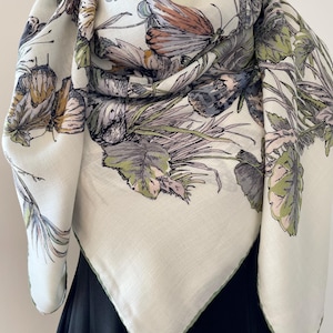 May include: A cream-coloured square scarf with a butterfly and botanical print. The scarf features butterflies in shades of brown, grey, and blue, with green and purple foliage. The edges are trimmed with a thin green border.