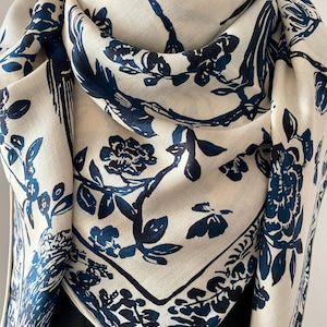 Puede incluir: Una bufanda color crema con un estampado floral azul oscuro. La bufanda está drapeada, mostrando el intrincado diseño de flores y hojas. Los bordes están bordeados con un ribete azul oscuro, agregando un toque de elegancia a este accesorio.