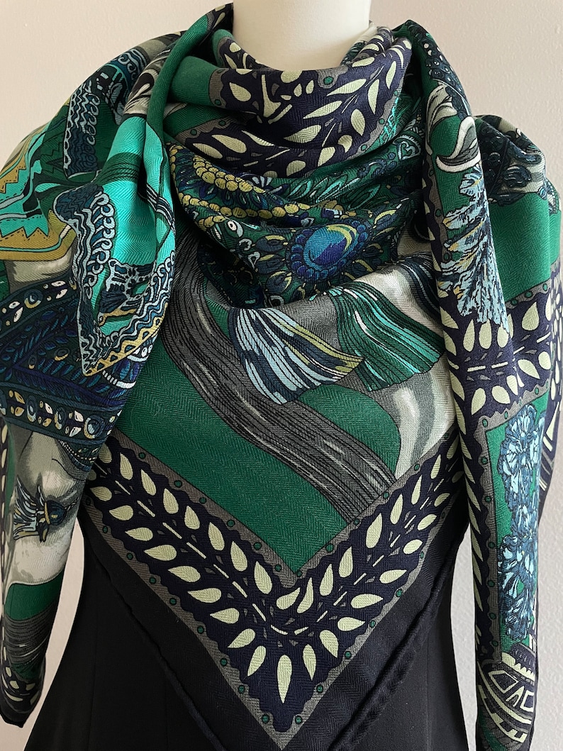 Peut inclure: Un carr&eacute; de soie orn&eacute; de motifs d&eacute;taill&eacute;s dans des tons de vert, turquoise, marine et gris. Le foulard pr&eacute;sente des motifs orn&eacute;s, notamment des &eacute;l&eacute;ments floraux et g&eacute;om&eacute;triques. Le foulard est drap&eacute; sur un v&ecirc;tement noir.