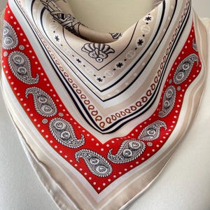 May include: A square silk scarf with a paisley and geometric design. The scarf features a red border with white polka dots and paisley motifs. The center is a cream color with black and brown accents. The scarf is folded and draped.